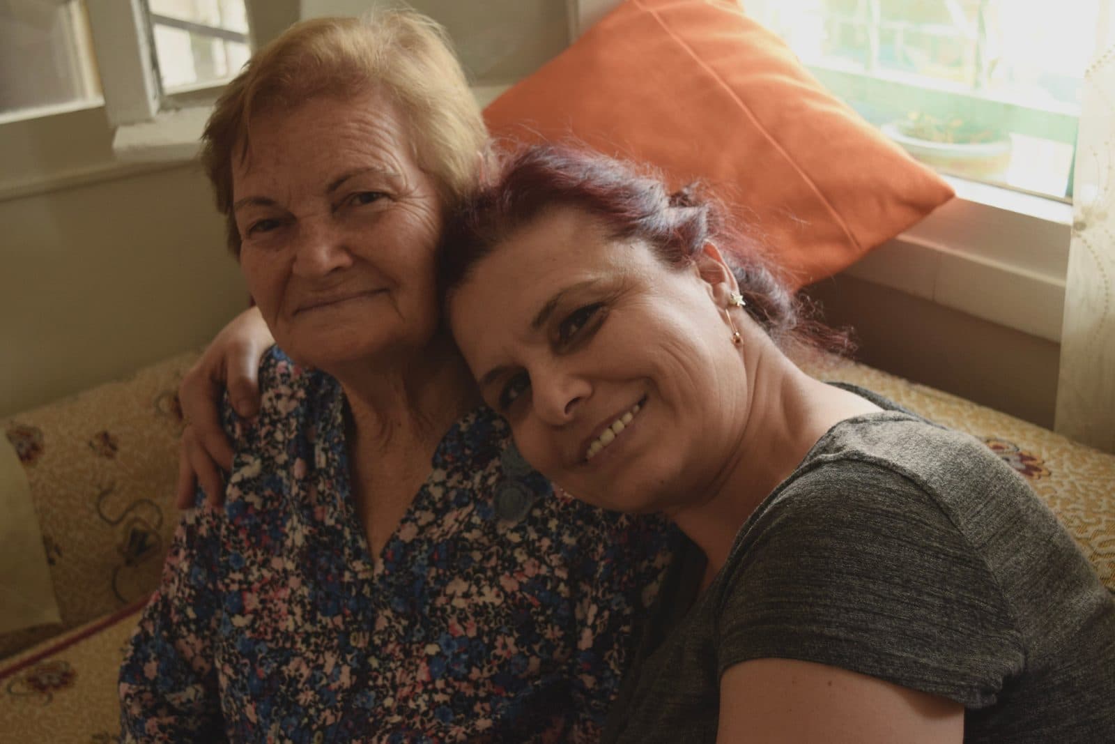 A woman sitting close to her mother at home, an arm around her shoulders.